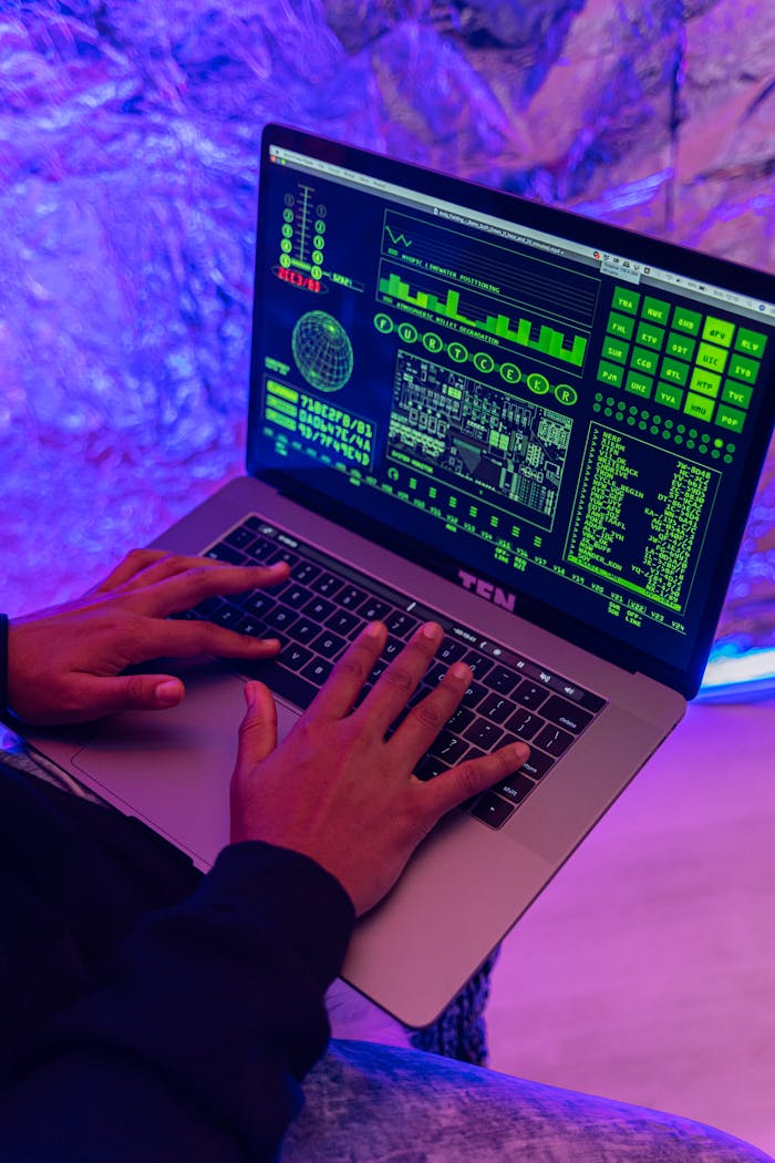 About Close-up of hands typing on a laptop displaying green code, indoors, purple lighting.
